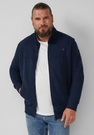 Felpa zip-up blu marina con colletto alto, in tessuto morbido e con un interno bianco a contrasto, abbinata a jeans azzurri chiari. Design semplice.
