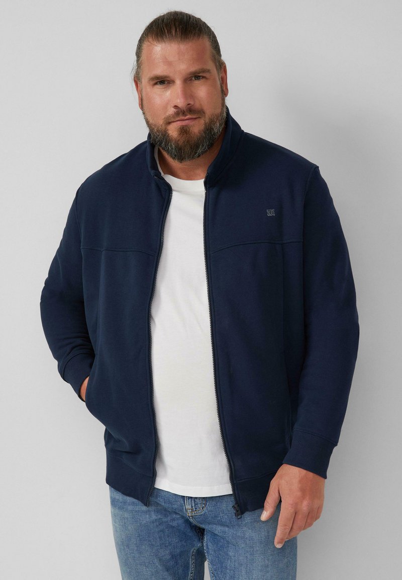 Felpa zip-up blu marina con colletto alto, in tessuto morbido e con un interno bianco a contrasto, abbinata a jeans azzurri chiari. Design semplice.