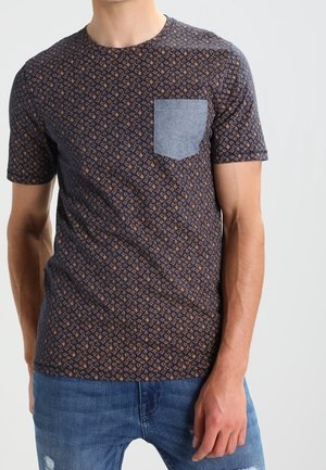 Mand iført en mørk mønstret kortærmet t-shirt med en ensfarvet lyseblå brystlomme og slidte blå jeans.