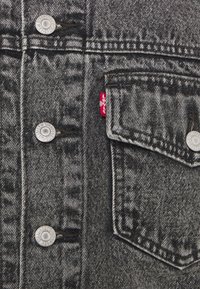 Giacca di denim nera con bottoni a pressione in metallo argentato, taschino sul petto e etichetta del marchio rossa, con una finitura strutturata e sbiadita.