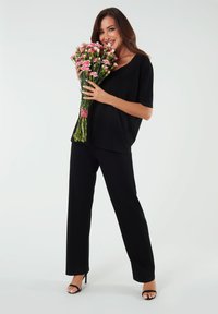 Czarna, luźno dopasowana bluzka i spodnie z gładkiego materiału. Modelka trzyma bukiet różowych goździków, ma na sobie czarne sandały na obcasie.