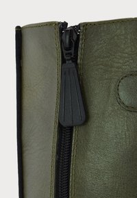 Gros plan sur une botte en cuir vert olive avec une fermeture éclair noire et une grande tirette noire.