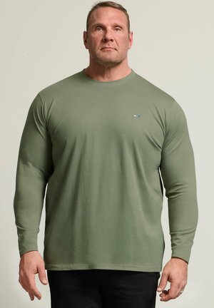 Mężczyzna z krótkimi włosami w prostym oliwkowo-zielonym długim rękawie, stojący na neutralnym tle, skierowany twarzą do przodu.