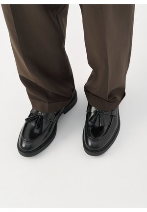 Zapatos de cuero negro con acabado brillante, detalle de borlas en la parte delantera y suela de goma, combinados con pantalones marrones de corte holgado.