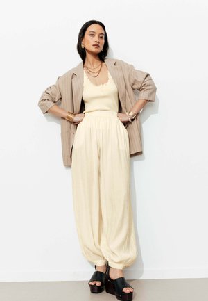 Femme appuyée contre un mur blanc portant un blazer beige à fines rayures, un top sans manches crème, un pantalon ample crème, des sandales à plateforme noires et des bijoux en or.