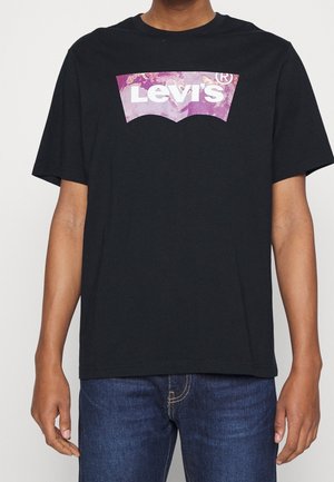 Personne portant un t-shirt Levi's noir avec un logo violet et blanc, associé à un jean en denim bleu.