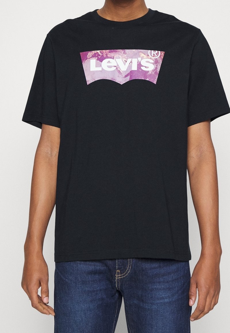 Personne portant un t-shirt Levi's noir avec un logo violet et blanc, associé à un jean en denim bleu.