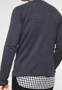 Maglione grigio scuro con un motivo a maglia testurizzato, scollo rotondo e maniche lunghe, indossato sopra una camicia a quadri bianca e nera.