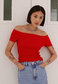 Top rosso scollato, realizzato in tessuto aderente, abbinato a una gonna di jeans sbiaditi a vita alta. Accessoriato con una collana in catena d'argento.