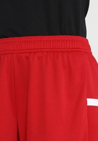 Pantalones cortos deportivos rojos con una tela texturada, cinturilla elástica y detalle de franjas horizontales blancas en el costado.