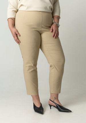 Beige, schmal zulaufende Hose aus Stretchstoff mit glatter Textur und mittelhohem Bund, kombiniert mit schwarzen spitzen Absatzschuhen.