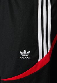 Pantaloni sportivi neri con strisce bianche sui lati, dettagli rossi e un logo bianco ricamato vicino all'orlo.