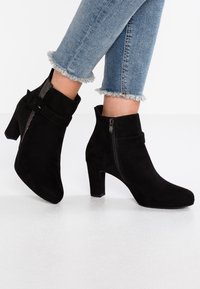 Zwarte suède enkellaarsjes met blokhak, voorzien van een ritssluiting aan de zijkant en een decoratieve gesp, gecombineerd met blauwe jeans met rafelige zoom.