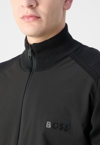 Black zip-up jacket with knitted collar, textured shoulder panels, and a subtle logo on the chest. Made from a smooth, lightweight fabric.