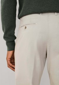 Pantalones beige con una textura suave y un bolsillo lateral con un detalle de botón de madera, combinados con un suéter de punto verde oscuro.