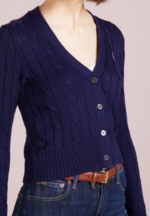 Frau trägt eine dunkelblaue, kabelgestrickte V-Ausschnitt-Strickjacke mit Knöpfen, in eine blaue Jeans gesteckt, die mit einem braunen Ledergürtel gesichert ist.