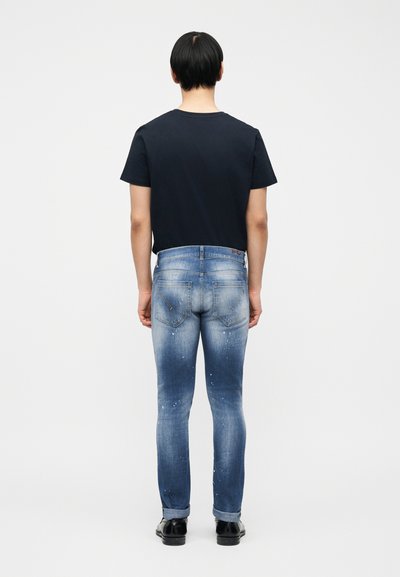 Άτομο που στέκεται με την πλάτη στο θεατή, φορώντας μαύρο μπλουζάκι, ξεθωριασμένο τζιν με πιτσιλιές μπογιάς και μαύρα παπούτσια, μπροστά σε απλό φόντο.