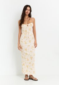Rochie maxi în crem deschis cu modele florale galbene, bretele subțiri, corset ajustat și detaliu plisat la talie, asociată cu sandale maro.