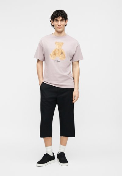 Νεαρός άνδρας με σγουρά μαλλιά που φοράει ανοιχτό μοβ μπλουζάκι με στάμπα αρκουδάκι, μαύρο κοντό παντελόνι, λευκές κάλτσες και μαύρα αθλητικά παπούτσια.