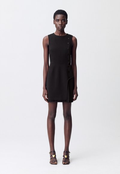 Una modella indossa un elegante abito nero con dettagli a bottoni laterali, abbinato a sandali neri con cinturini, su uno sfondo minimalista.