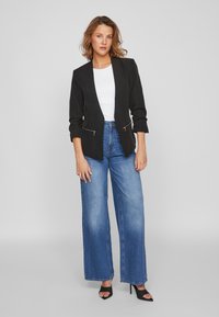 Blazer preto com bolsos com fecho, combinado com jeans azuis de perna larga e alta na cintura. Modelo a usar tacões pretos de ponta aberta, com uma blusa branca por baixo.