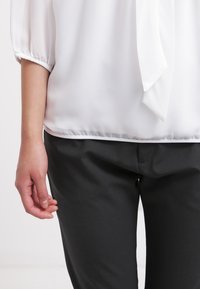 Left hand resting beside black pants, wearing a white blouse with three-quarter sleeves and a necktie detail.