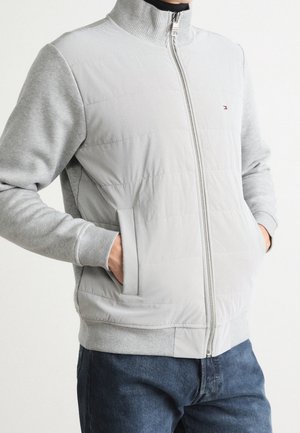 Mand iført lys grå lynlåstrøje med quiltet front og strikkede ærmer, hænder i lommerne, kombineret med blå jeans.