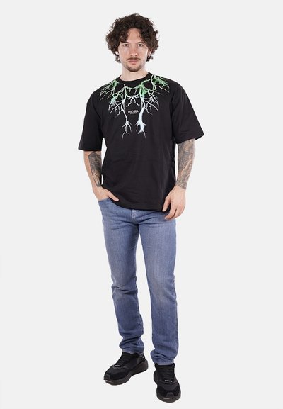 Maglietta nera con grafica di un fulmine verde sul collo; maniche corte; abbinata a jeans azzurri chiari e sneakers nere.