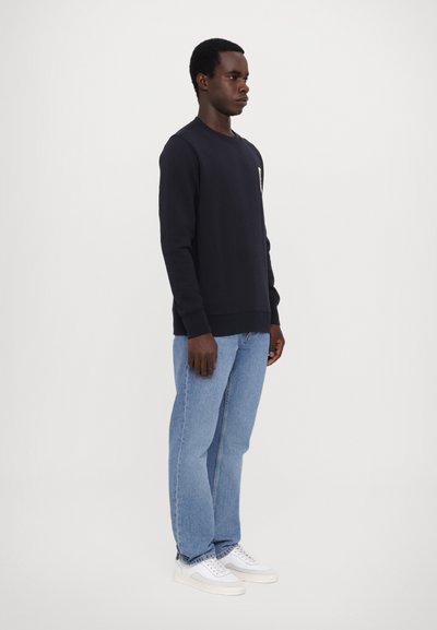 Tummansininen collegepaita pitkillä hihoilla, pyöreällä kaula-aukolla ja joustinsuurilla hihoilla, yhdistettynä vaaleansinisiin farkkuihin ja valkoisiin tennareihin.