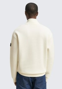 Witte gebreide pullover met hoge kraag en geribbelde manchetten, voorzien van een subtiel patroon. De achterkant laat een minimale merkaanduiding op de mouw zien.