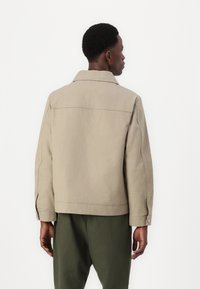 Beige cotton jacket with a collar, back yoke detail, and side slits. Features long sleeves with buttoned cuffs. Worn over olive green trousers.
