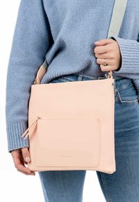 Růžová, hladká syntetická krosbody taška s přední kapsou na zip. Nastavitelný popruh a minimalistický design. Nosená s modrým svetr a džínami.