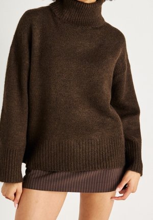 Persona che indossa un maglione marrone a collo alto in maglia grossa e una gonna corta marrone a righe sottili, mani rilassate lungo i fianchi.