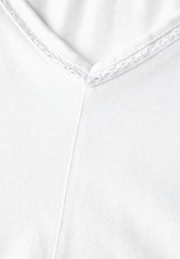T-shirt bianco in cotone con scollatura a V e dettagli in pizzo rifiniti sul bordo, texture liscia e cuciture senza cuciture lungo il tessuto.