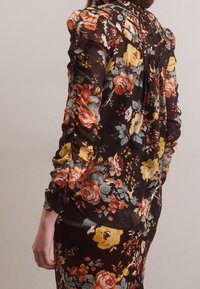 Blouse fleurie en tissu noir avec des fleurs orange, jaunes et vertes. Détails plissés au niveau de l'encolure et manches froncées ajoutent de la texture.