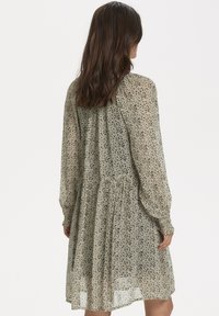 Robe à manches longues en tulle beige clair avec un motif abstrait noir, présentant une taille froncée et une jupe fluide.