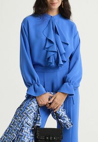 Blusa blu a maniche lunghe con dettagli arricciati, chiusura con bottoni e polsini aderenti; abbinata a una sciarpa fantasia e una piccola borsa nera.