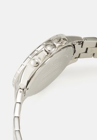 Montre en acier inoxydable argenté avec un design chronographe, textures brossées et polies, et boutons proéminents sur le côté.