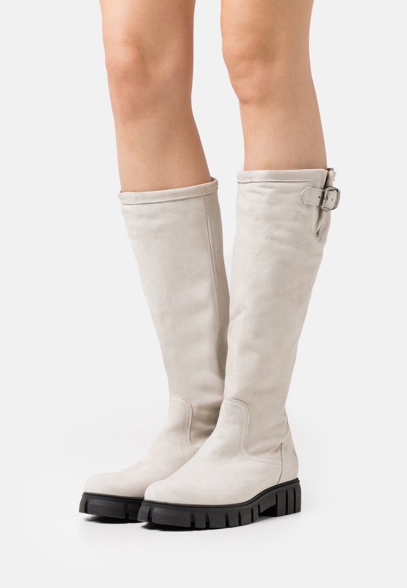 Femme portant des bottes en cuir beige clair arrivant au genou avec des semelles noires épaisses et une boucle sur la botte droite, se tenant sur un fond blanc.