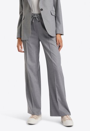 Kvinde iført grå bukser med vide ben og snoretræk i taljen, matchende grå blazer, hvid top og beige sneakers, stående mod en hvid baggrund.