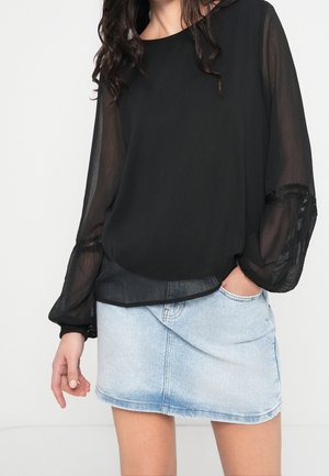 Femme portant un chemisier noir transparent à manches longues rentré dans une mini-jupe en jean bleu clair, une main dans la poche de la jupe.