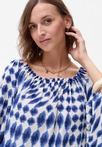 Niebiesko-biała bluzka w wzory z marszczonym dekoltem, wykonana z lekkiego materiału. Modelka uzupełniła strój złotym naszyjnikiem.