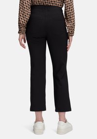 Vrouw in zwarte cropped broek, met een patroon op een lange mouwen blouse en witte sneakers, staand met de rug naar de camera op een witte achtergrond.