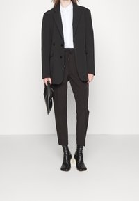 Blazer noir à double boutonnage, chemise blanche à col, pantalon noir à cordon de serrage et bottines noires, tenant une pochette noire.