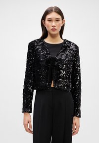 Μαύρο σακάκι με παγιέτες, κομμένο σε κοντό μήκος, με υφή, μακριά μανίκια και κλεισιμο με φιόγκο μπροστά, φορεμένο πάνω από ένα μαύρο τοπ.