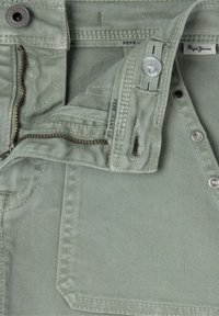 Zelené bavlnené džínsy so zapínaním na zips a gombík, s textúrovaným povrchom a zosilneným šitím. Značka viditeľná vo vnútri pásu.