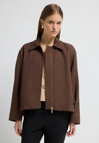 Brun cropped jakke med en struktureret krave og lynlås foran, fremstillet i et tekstureret stof med brede ærmer. Bæret over en beige top og sorte bukser.