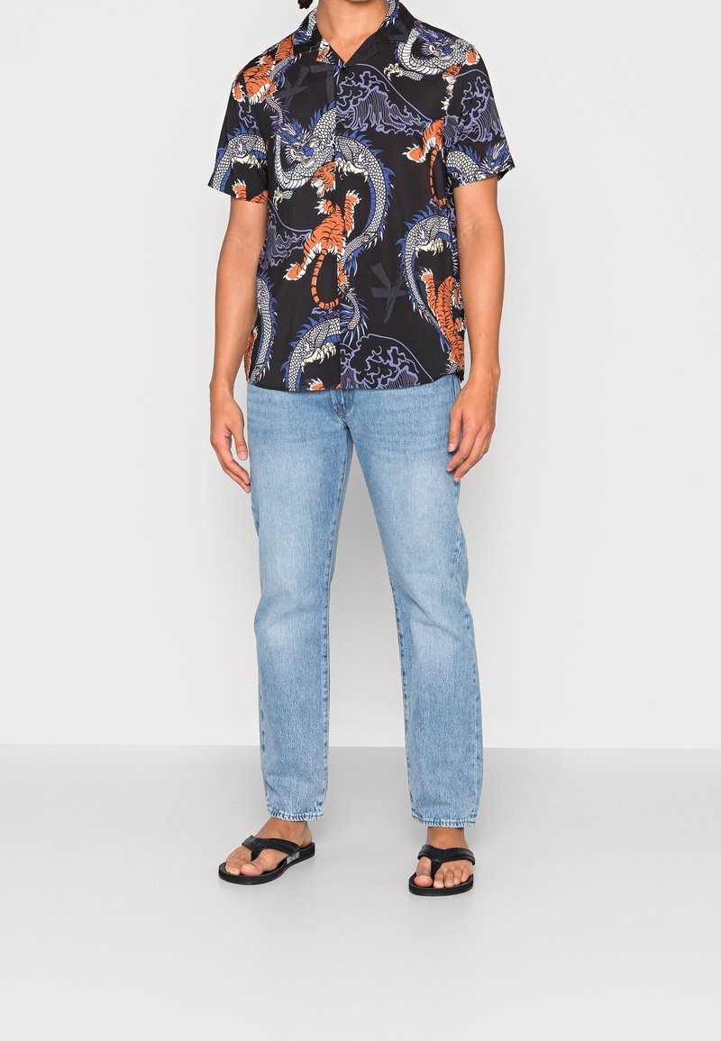 Lyhythihainen musta paita, jossa on värikkäitä lohikäärme- ja tiikerikuvioita, yhdistettynä vaaleansinisiin farkkuihin ja mustiin varvastossuihin neutraalilla taustalla.