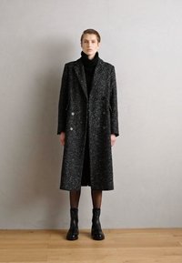Langer, doppelt knöpfbarer Mantel aus strukturiertem schwarzen und grauen Stoff, mit Taschen auf der Vorderseite und dunklen Knöpfen, getragen über einem schwarzen Rollkragenpullover.