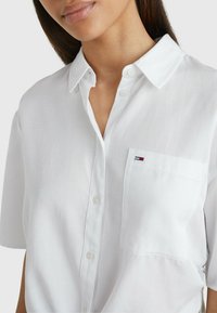 Camicia bianca a maniche corte con colletto a punta, dotata di taschino sul petto e un piccolo logo decorativo. Realizzata in tessuto leggero.